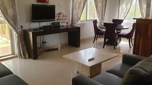 Een woonkamer met een bank, een salontafel, een televisie, een bureau en een eethoek met stoelen.