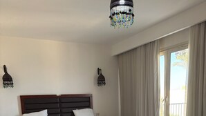 A bedroom with a bed, bedside tables, a bench, a chandelier, and a view through a sliding door. - Carlton Luxury Resort (Sharm El Sheikh)