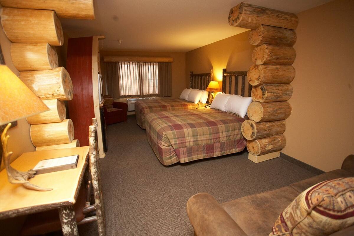 A hotel room with two beds, a wooden log wall, a desk with a lamp, and a window with curtains.
