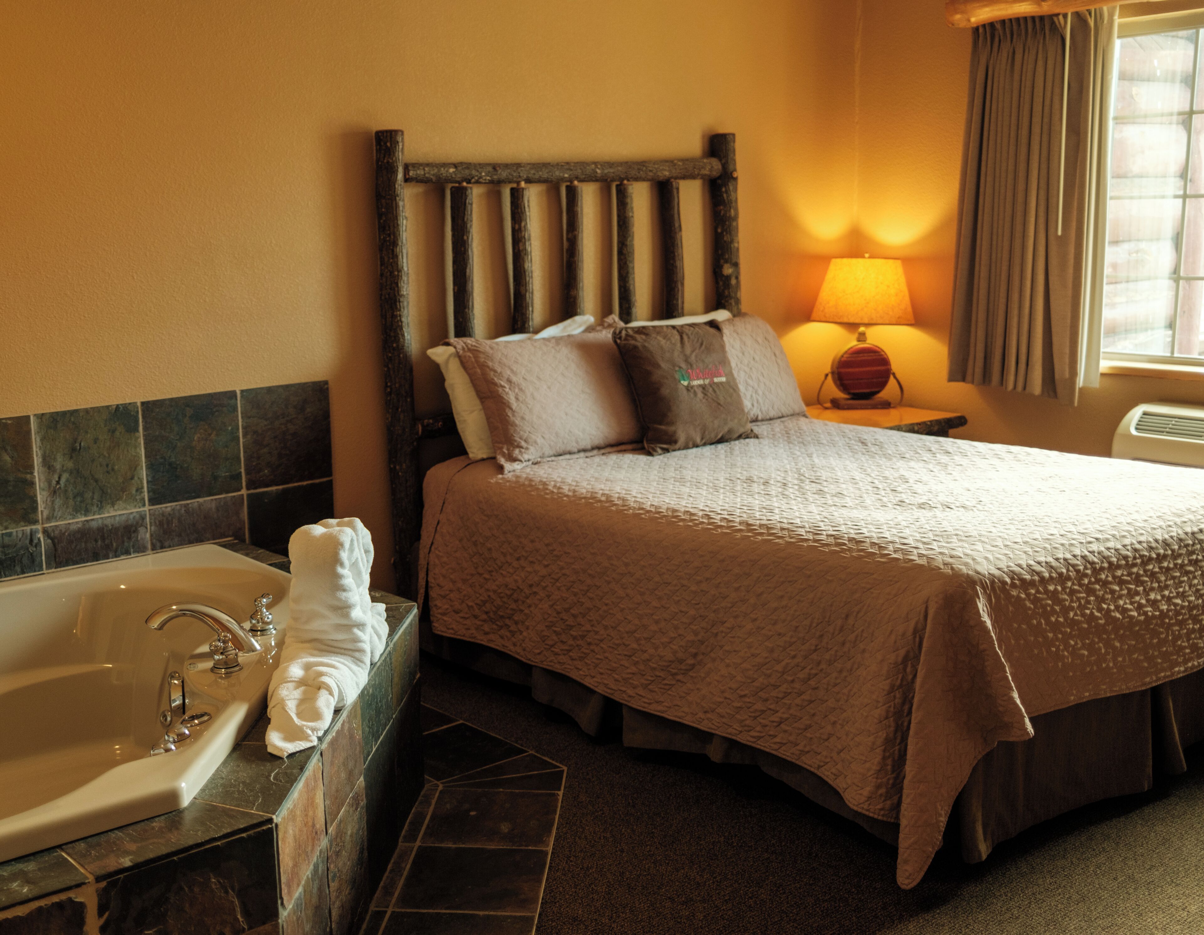 A hotel room with a bed, a nightstand, a lamp, a bathroom with a bathtub, and a window with curtains.
