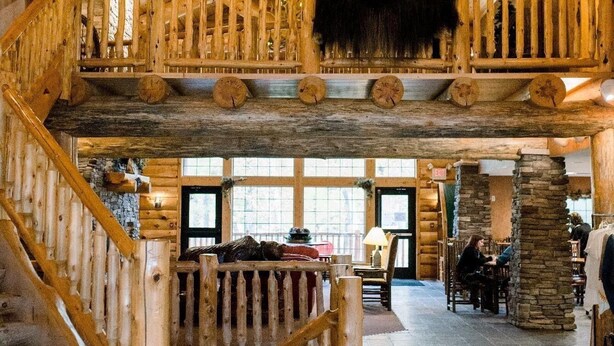 A spacious log cabin interior with a central staircase, stone pillars, and a rustic wooden ceiling. - Whitefish Lodge and Suites (Crosslake)