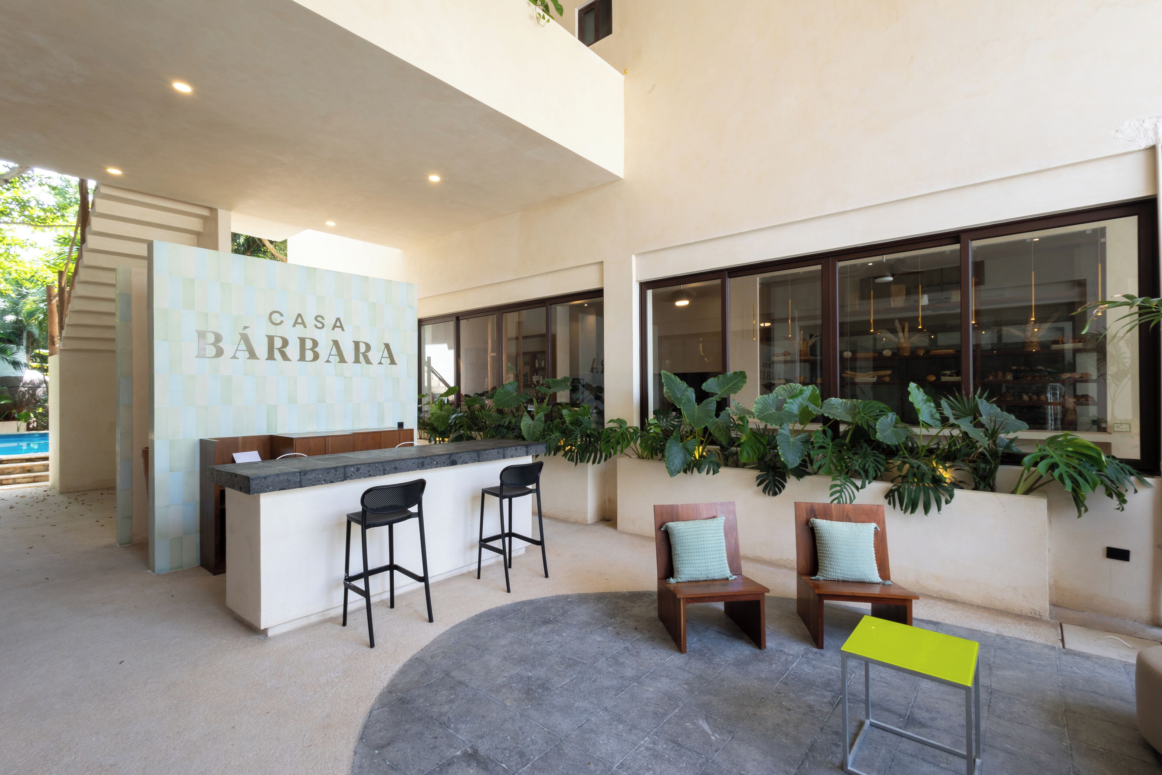 A modern bar area with a sign reading "Casa Bárbara," two bar stools, and a seating area with two chairs and a small table.
