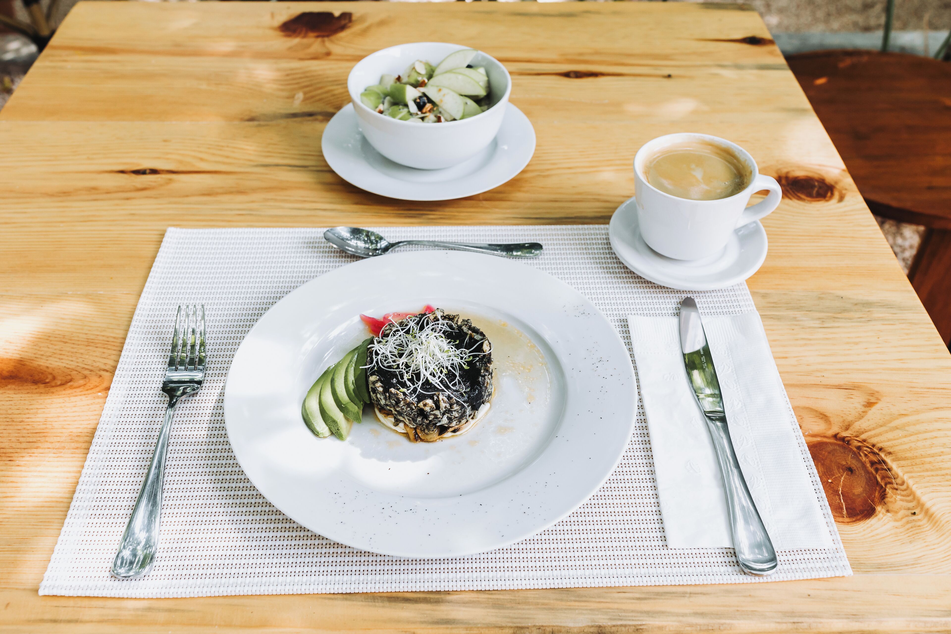 A wooden table with a white placemat, a white plate with a food item, a fork, a spoon, a knife, a white cup with a beverage, and a white bowl with a salad.