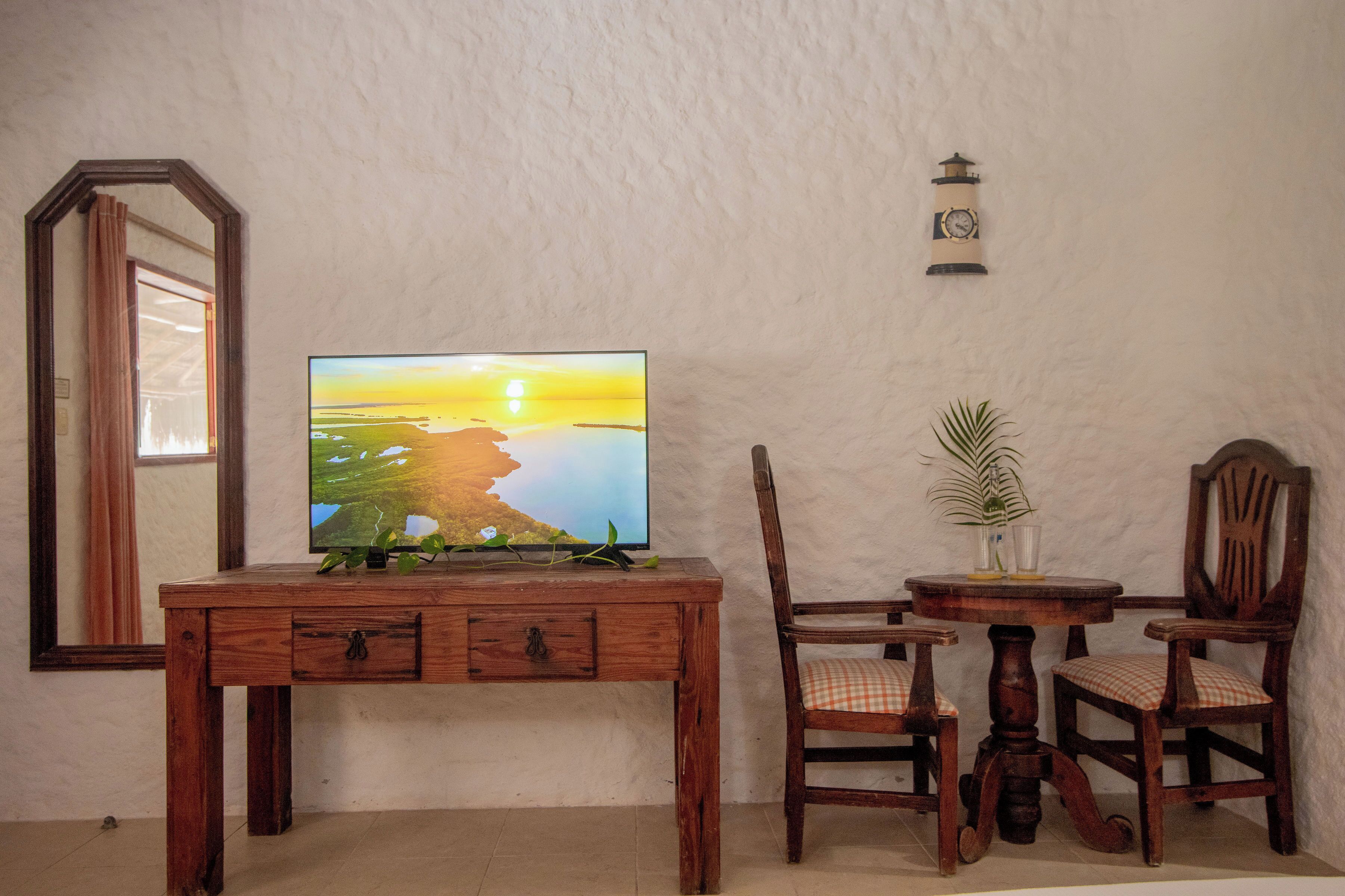 A wooden desk with a flat-screen TV, two chairs, a small table with a plant, and a wall-mounted clock.