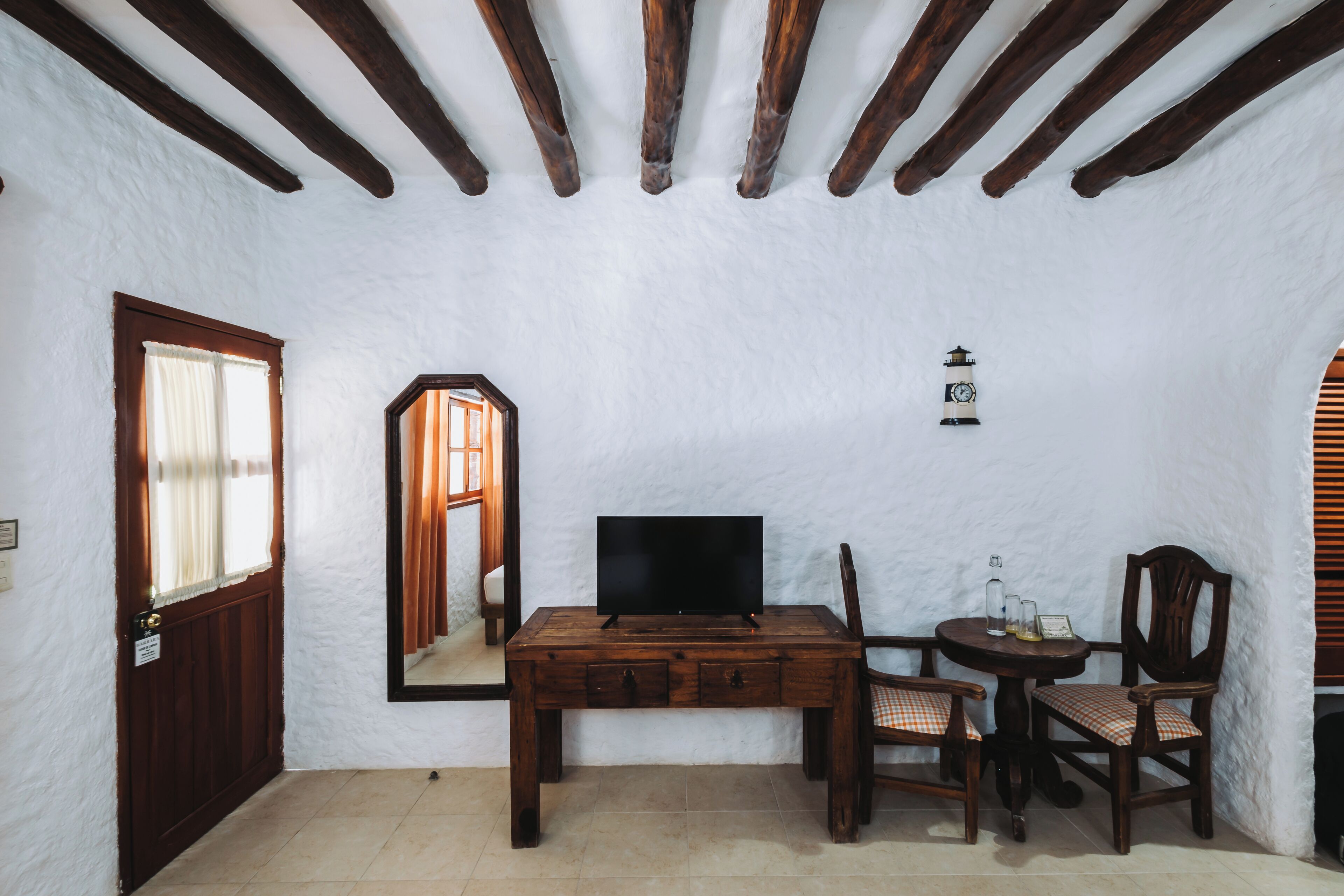 Una habitación con un escritorio de madera, televisor, silla, una mesita con una botella y vasos, y un espejo.