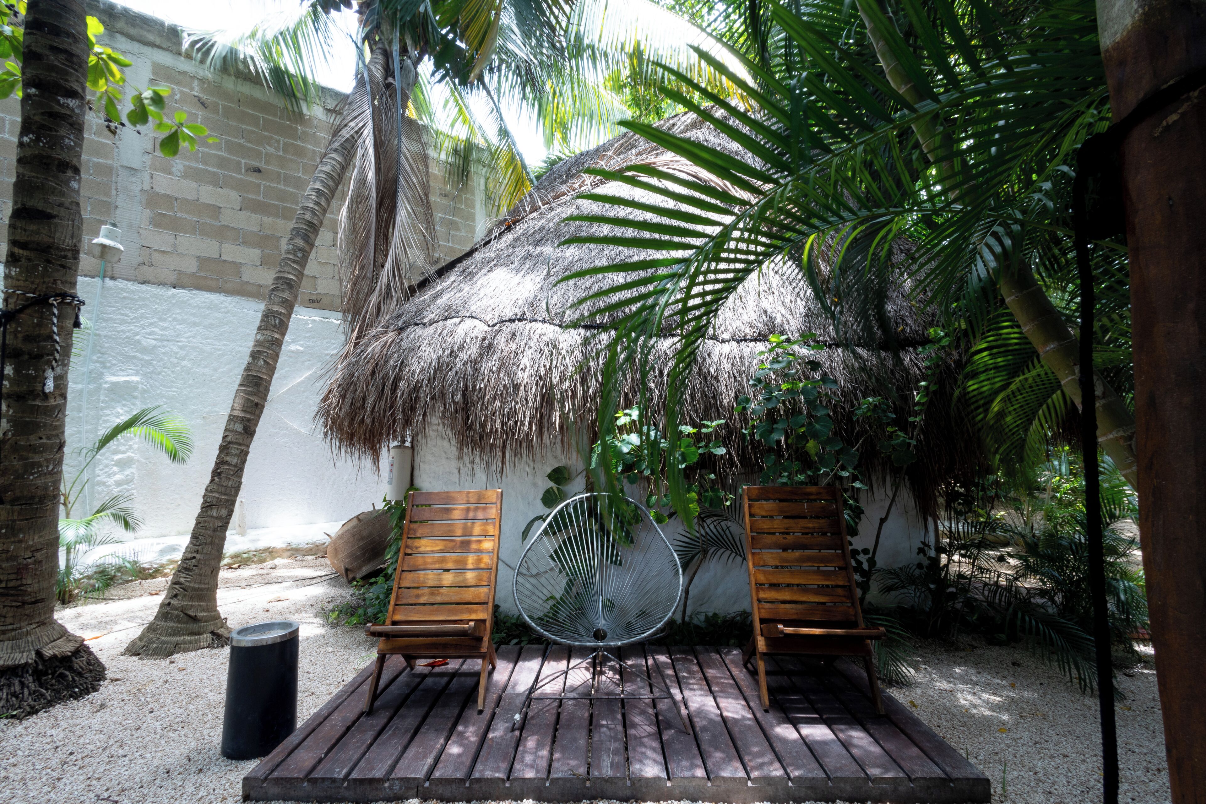 A wooden deck with two wooden chairs and a metal fan, surrounded by palm trees and a thatched roof structure.