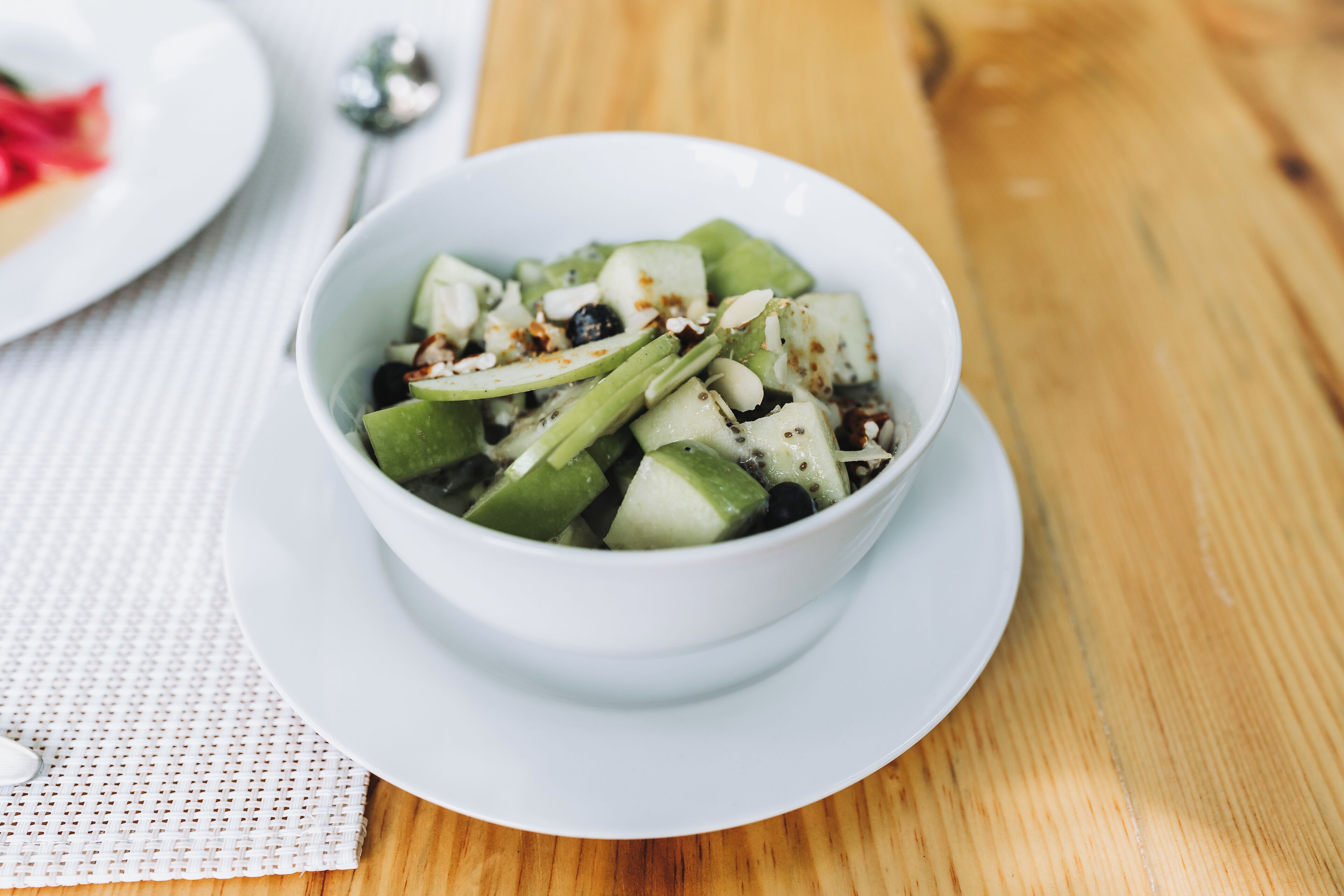 A white bowl of sliced green fruit salad on a white plate, placed on a wooden table.
