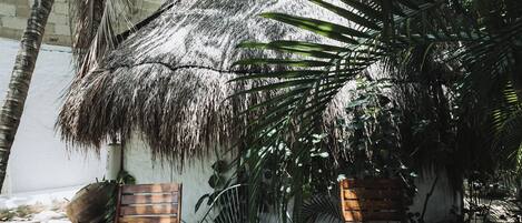 A thatched-roof outdoor seating area with two wooden chairs and a fan, surrounded by palm trees and a brick wall.