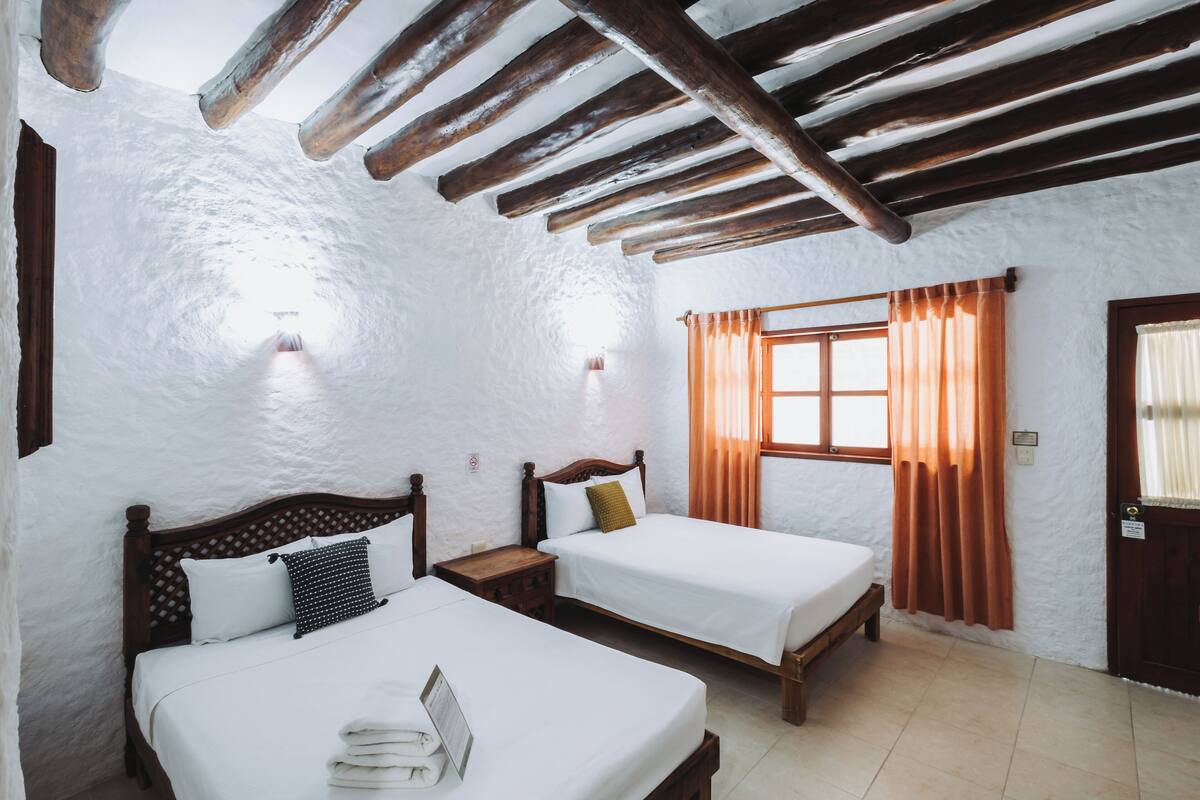 Ein Zimmer mit zwei Betten, Holz-Kopfteilen, weißen Wänden, einer Holzbalkendecke, einem Fenster mit Vorhängen und einer Tür.