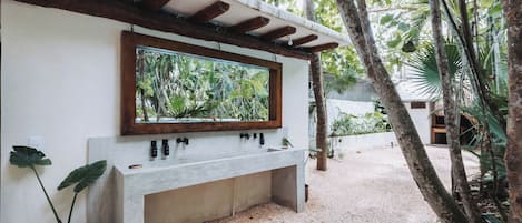 A modern outdoor bathroom with a concrete sink, wooden beams, and a large mirror reflecting the surrounding greenery.