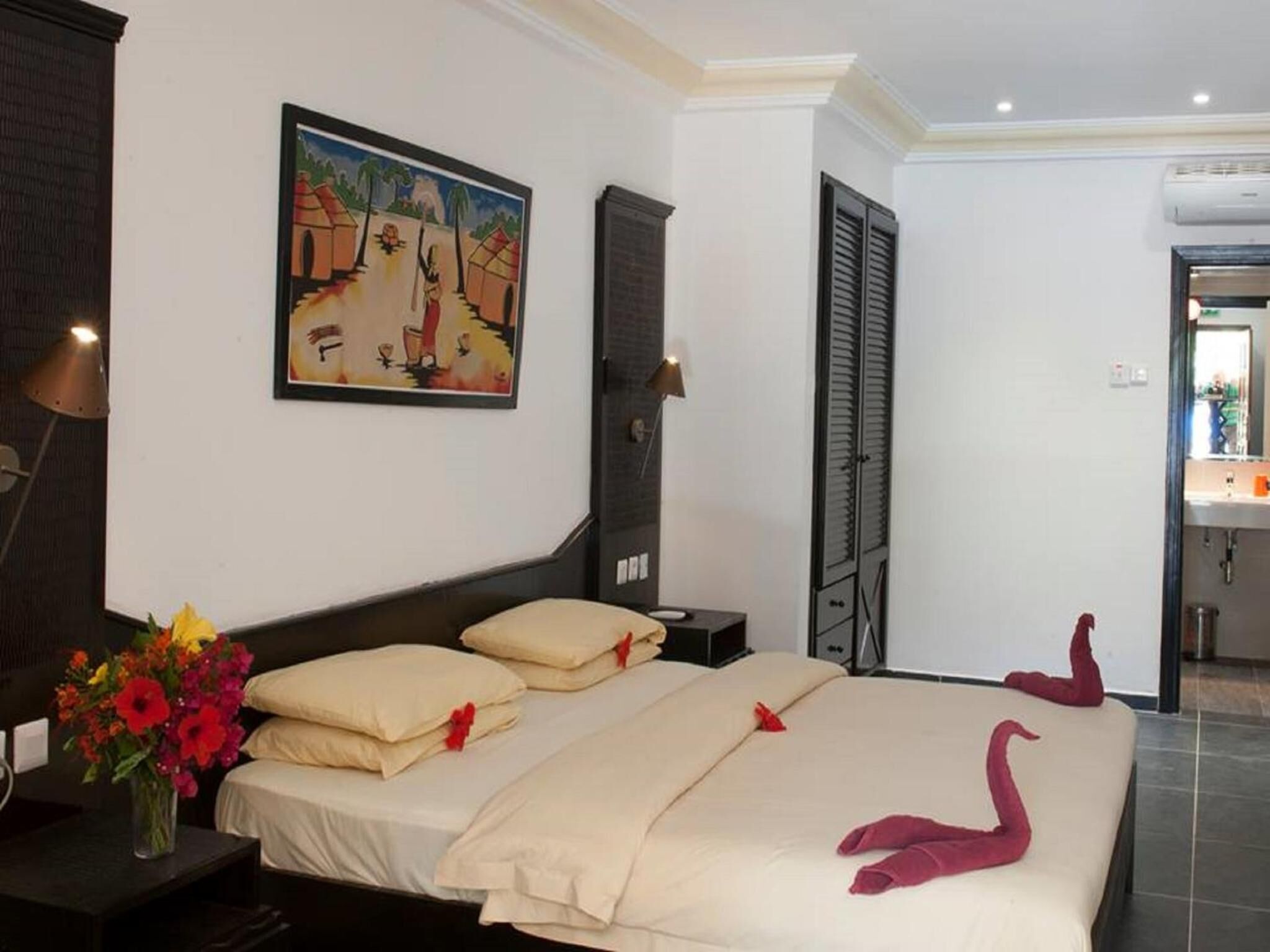 Une chambre d’hôtel avec un lit, une table de chevet, une lampe, un tableau et une salle de bain visible à travers une porte ouverte.