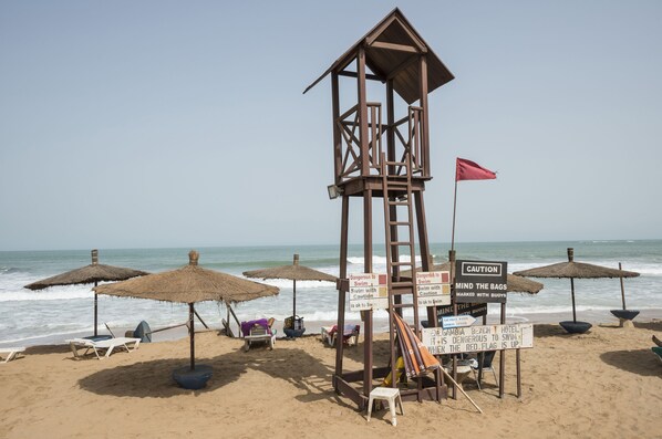 Una playa con una torre de salvavidas, sombrillas, sillas de descanso y señales de precaución.