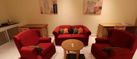 A living room with a red sofa set, a wooden coffee table, and two armchairs. There are two paintings on the wall and a small table with a chair.