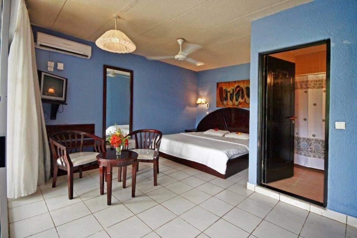 Ein Hotelzimmer mit einem Bett, zwei Stühlen, einem kleinen Tisch mit Blumen, einem Fernseher und einem Deckenventilator.