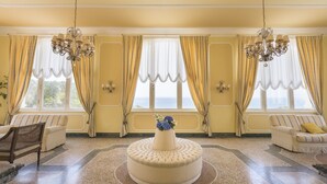 A room with a round ottoman, two sofas, a chandelier, and large windows with curtains. - Grand Hotel Villa Parisi (Rosignano Marittimo)