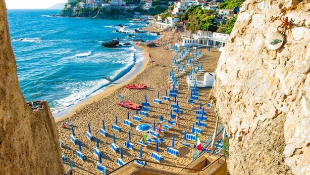 A beach with rows of blue umbrellas and chairs, a rocky cliff, and a view of the sea and buildings in the background. - Grand Hotel Villa Parisi (Rosignano Marittimo)