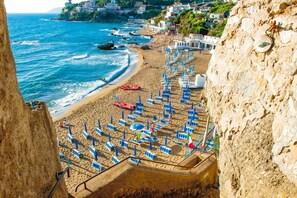 A beach with rows of blue umbrellas and chairs, a rocky cliff, and a view of the sea and buildings in the background. - Grand Hotel Villa Parisi (Rosignano Marittimo)