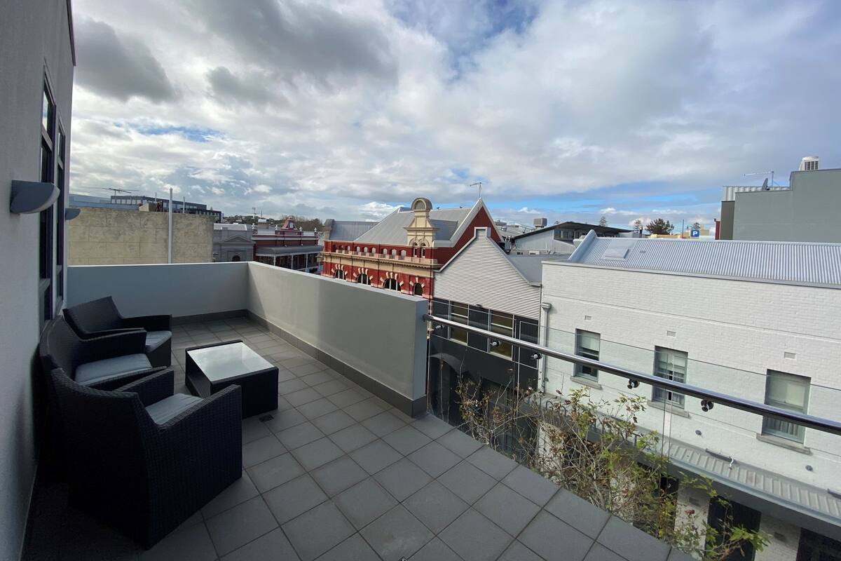 A rooftop terrace with outdoor furniture, a view of historic buildings, and a clear sky.
