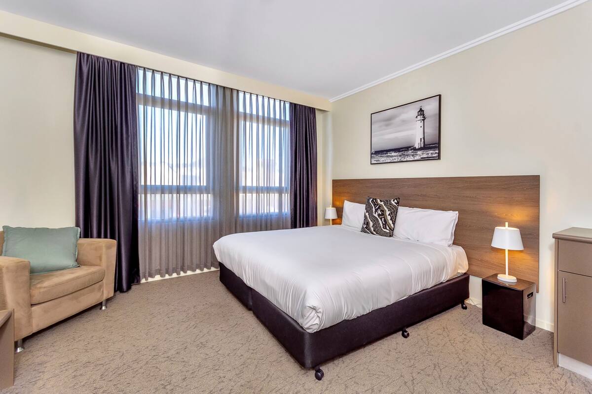 A hotel room with a large bed, a nightstand with a lamp, a chair, and a painting of a lighthouse.