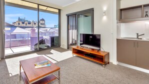 A modern living room with a flat-screen TV, a wooden coffee table, and a large sliding glass door leading to a balcony with a city view. - New Haus Managed by Hougoumont (Fremantle)