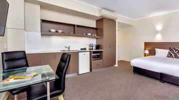 Una habitación de hotel compacta con kitchenette, una cama, una mesa de vidrio y una silla.