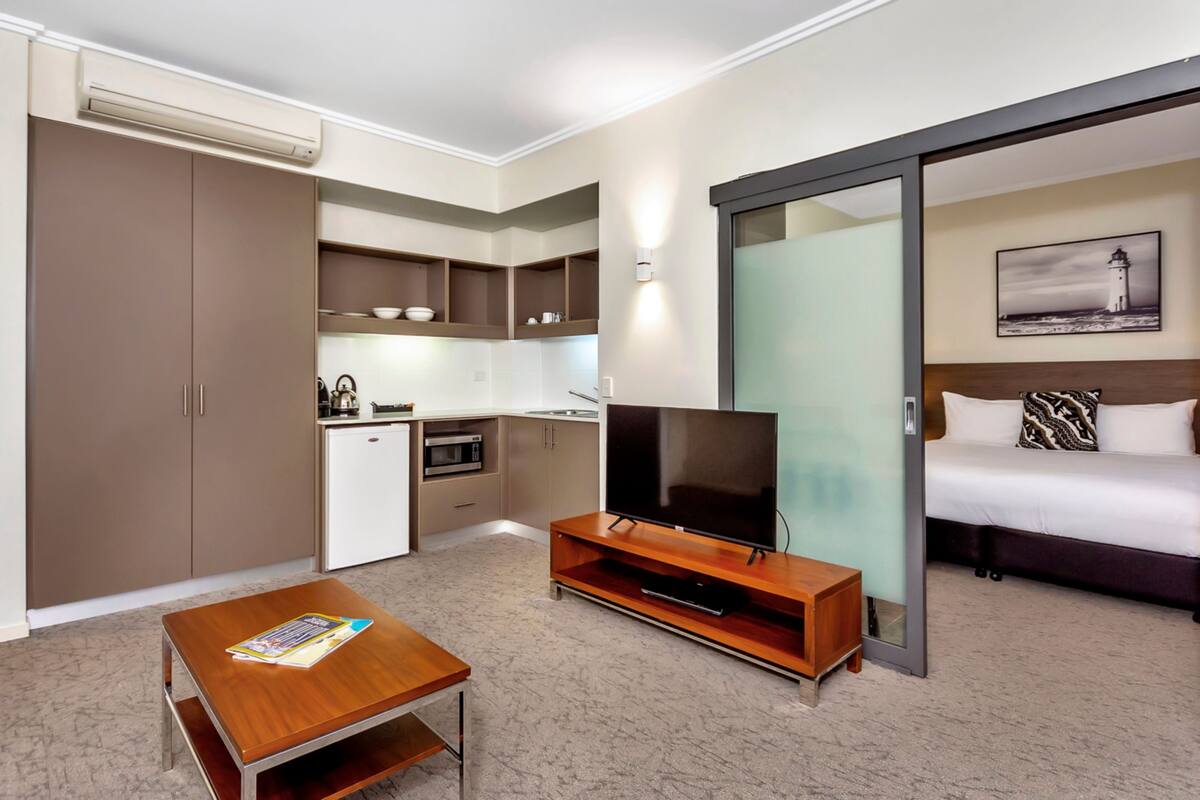 A compact hotel room with a kitchenette, a flat-screen TV on a wooden stand, a bed with patterned pillows, and a sliding glass door leading to a balcony.
