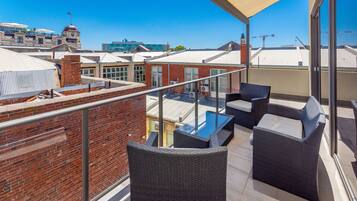 A rooftop terrace with wicker furniture, a glass table, and a view of surrounding buildings.