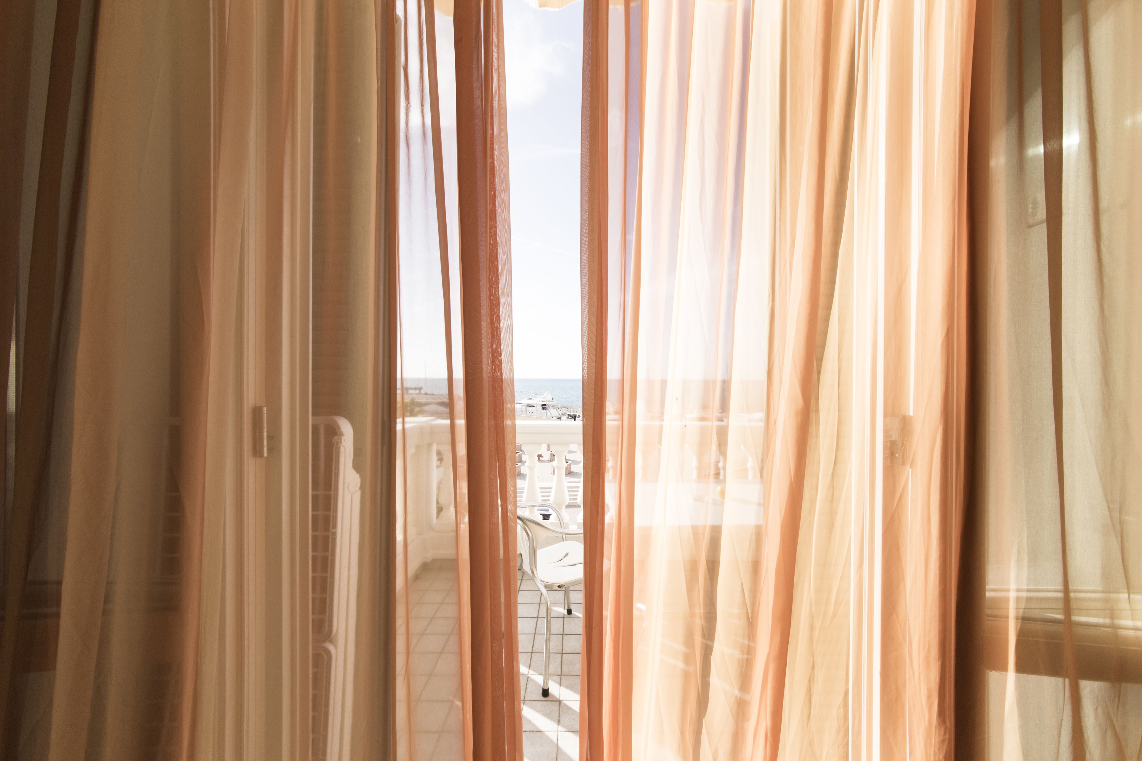 Vista a través de cortinas translúcidas hacia un balcón con sillas blancas y piso de baldosas.