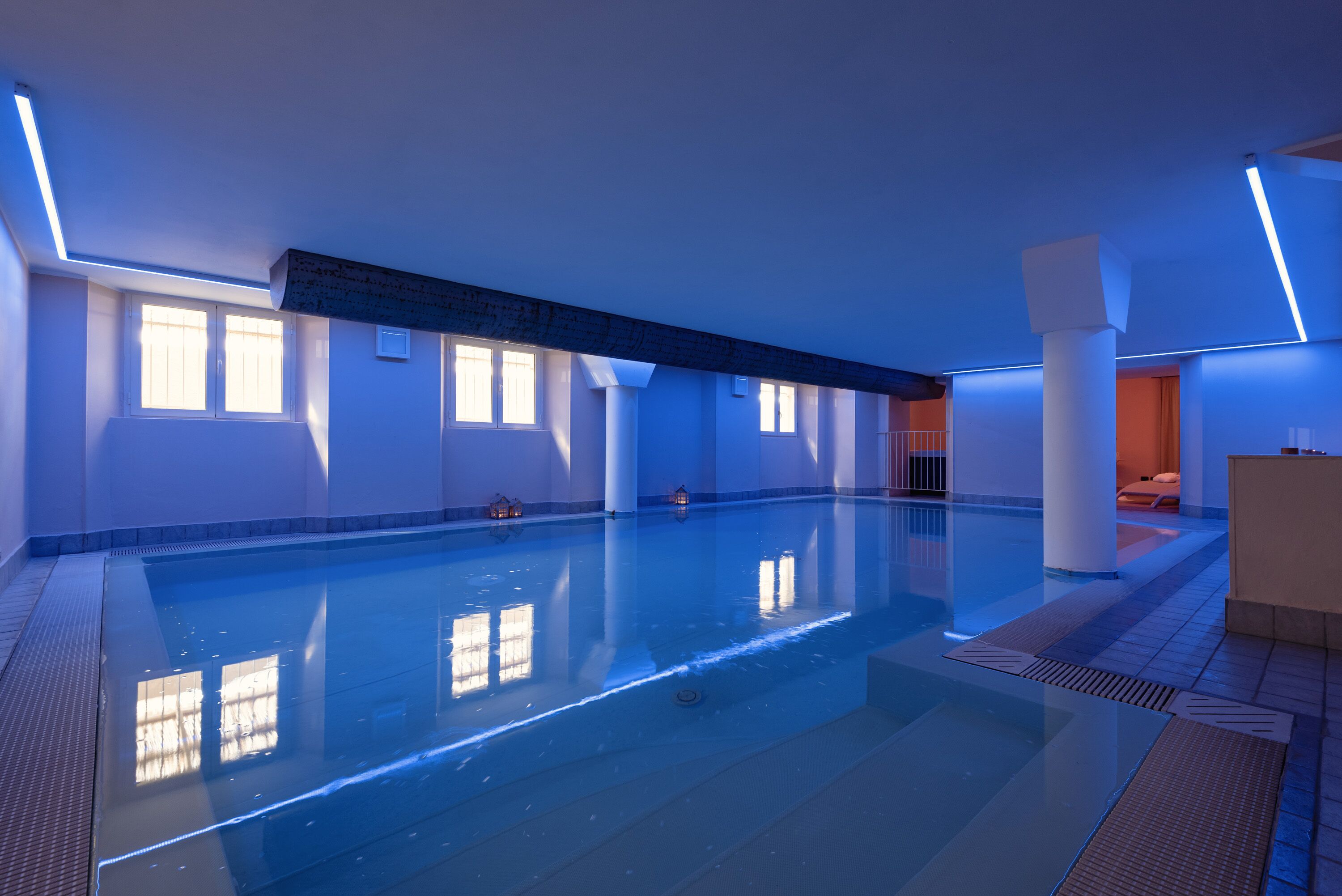 A large indoor swimming pool with clear water, surrounded by white walls and columns, and natural light coming through windows.