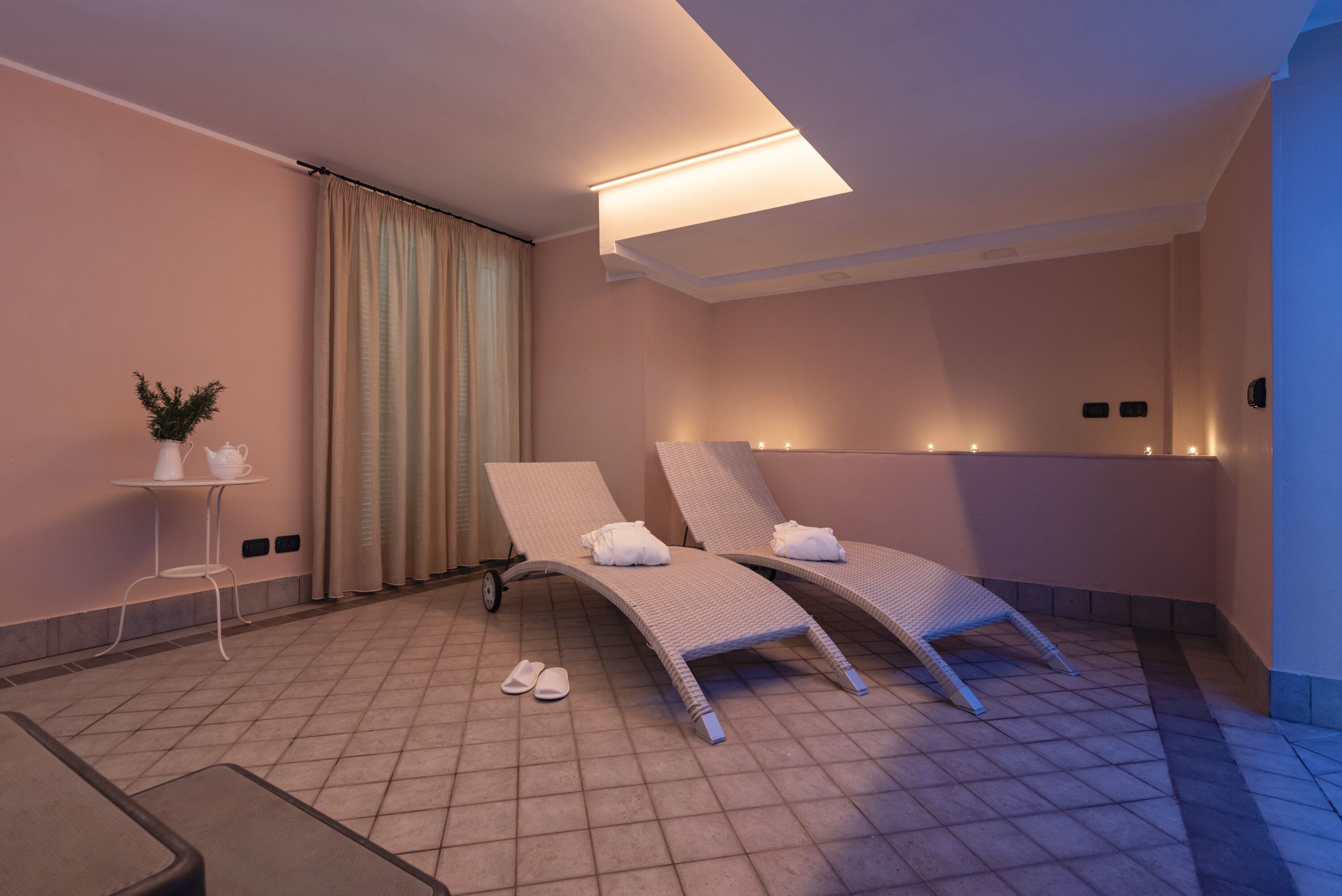 A spa room with two reclining chairs, a small table with a plant, and a pair of slippers on the floor.