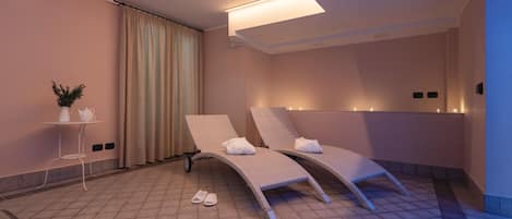 A spa room with two reclining chairs, a small table with a plant, and a pair of slippers on the floor.