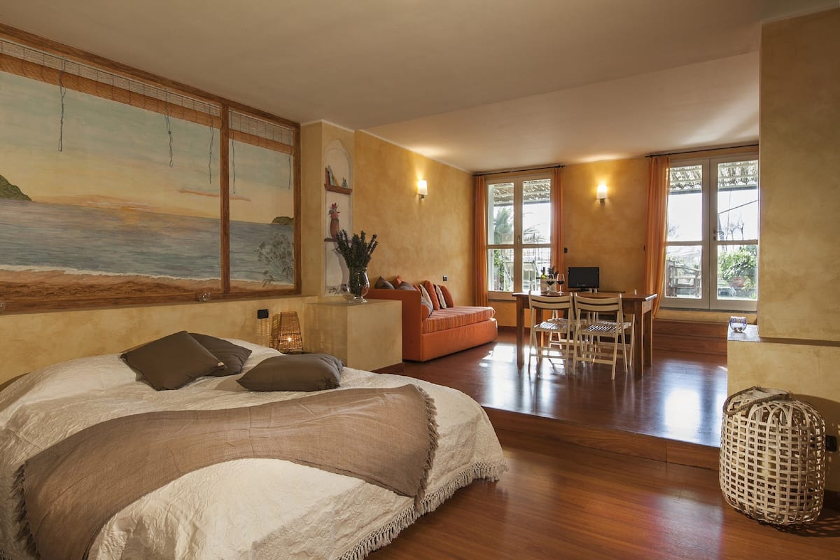 A bedroom with a large bed, a desk, and a sofa. There is a painting on the wall and a window with curtains.