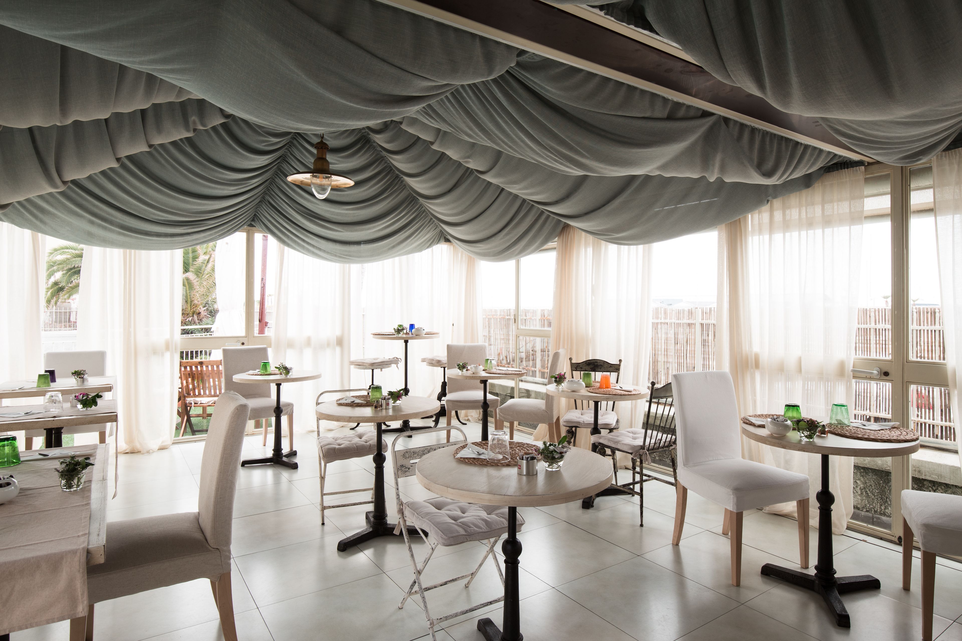 A spacious dining area with round tables and white chairs, large windows with sheer curtains, and a view of palm trees outside.