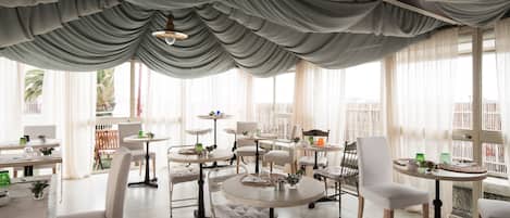 A spacious dining area with round tables and white chairs, large windows with sheer curtains, and a view of palm trees outside.