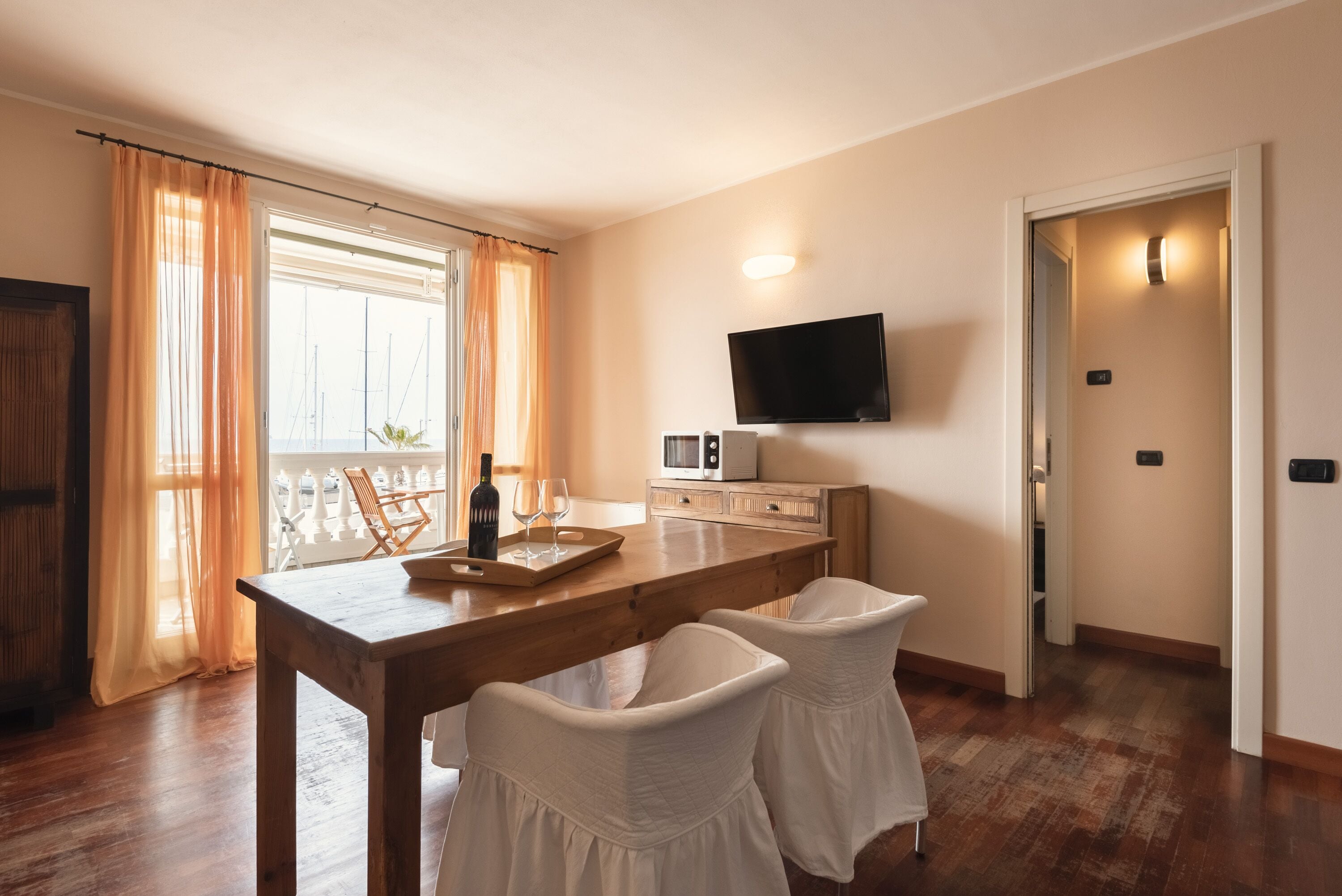 Un comedor con una mesa de madera, dos sillas blancas, un televisor de pantalla plana en la pared y una ventana con cortinas.