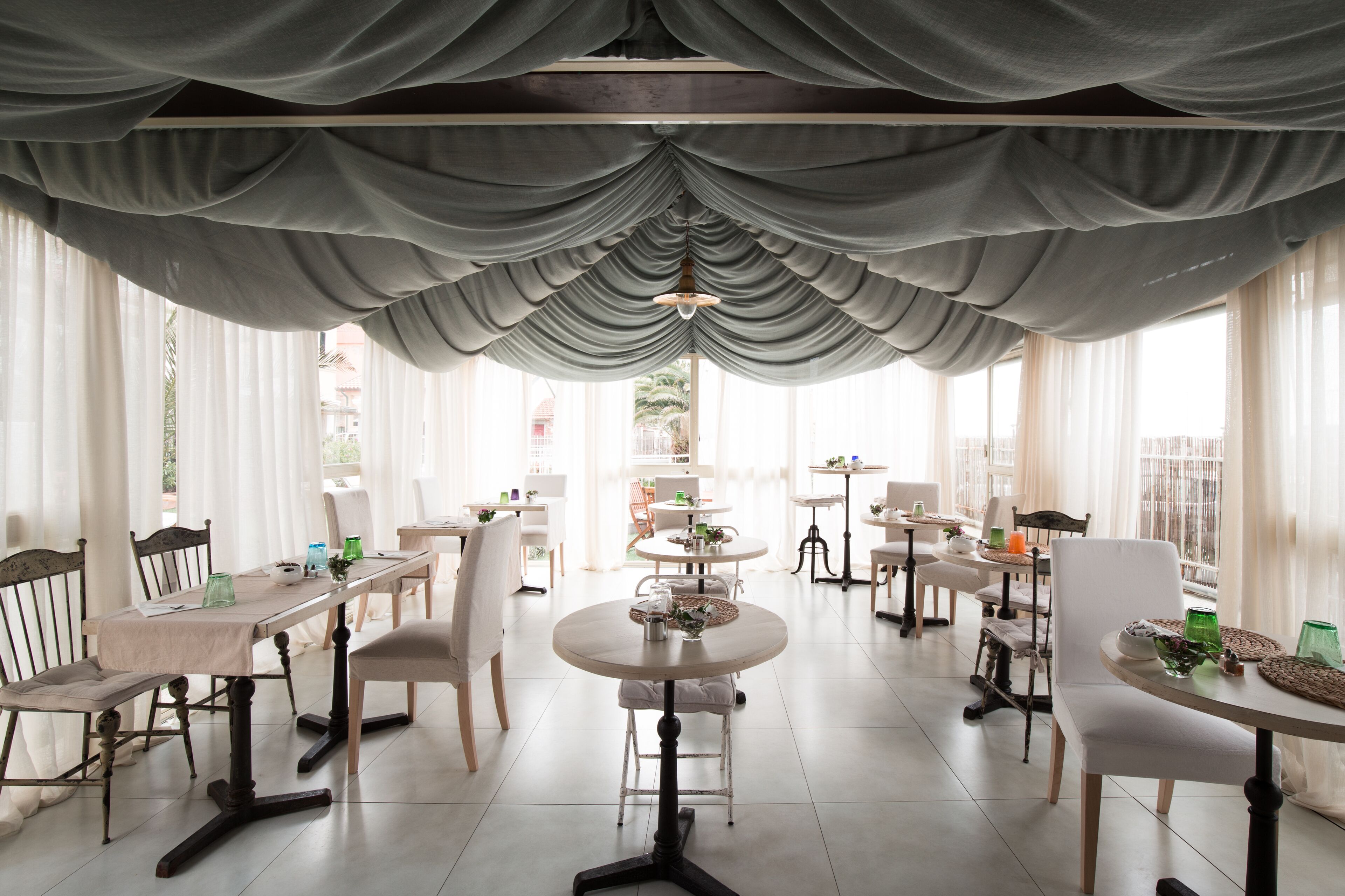 A dining area with tables and chairs, draped curtains, and large windows.