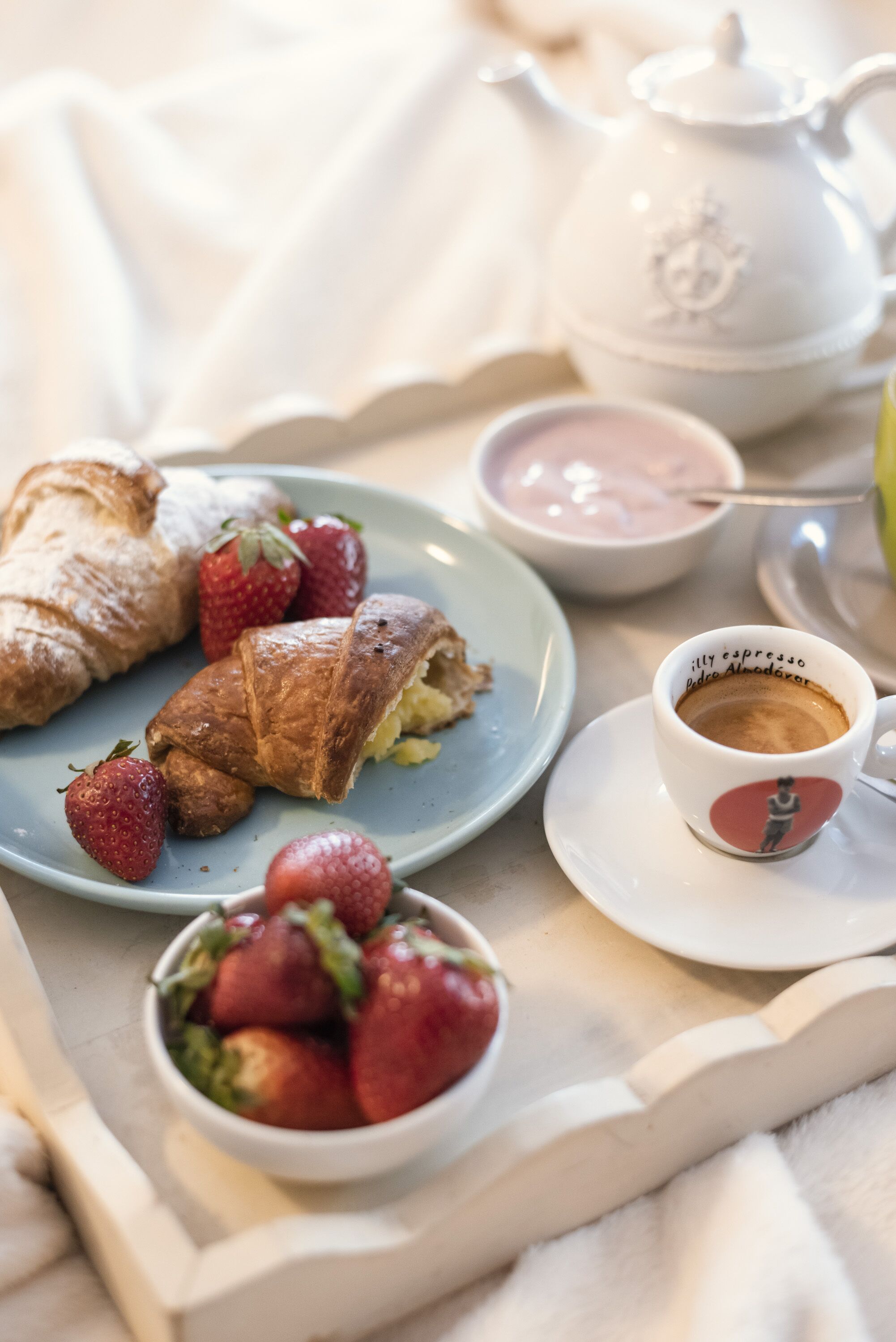 Aamiaispöytä, jossa on croissantteja, mansikoita, teekannu ja kuppi kahvia.