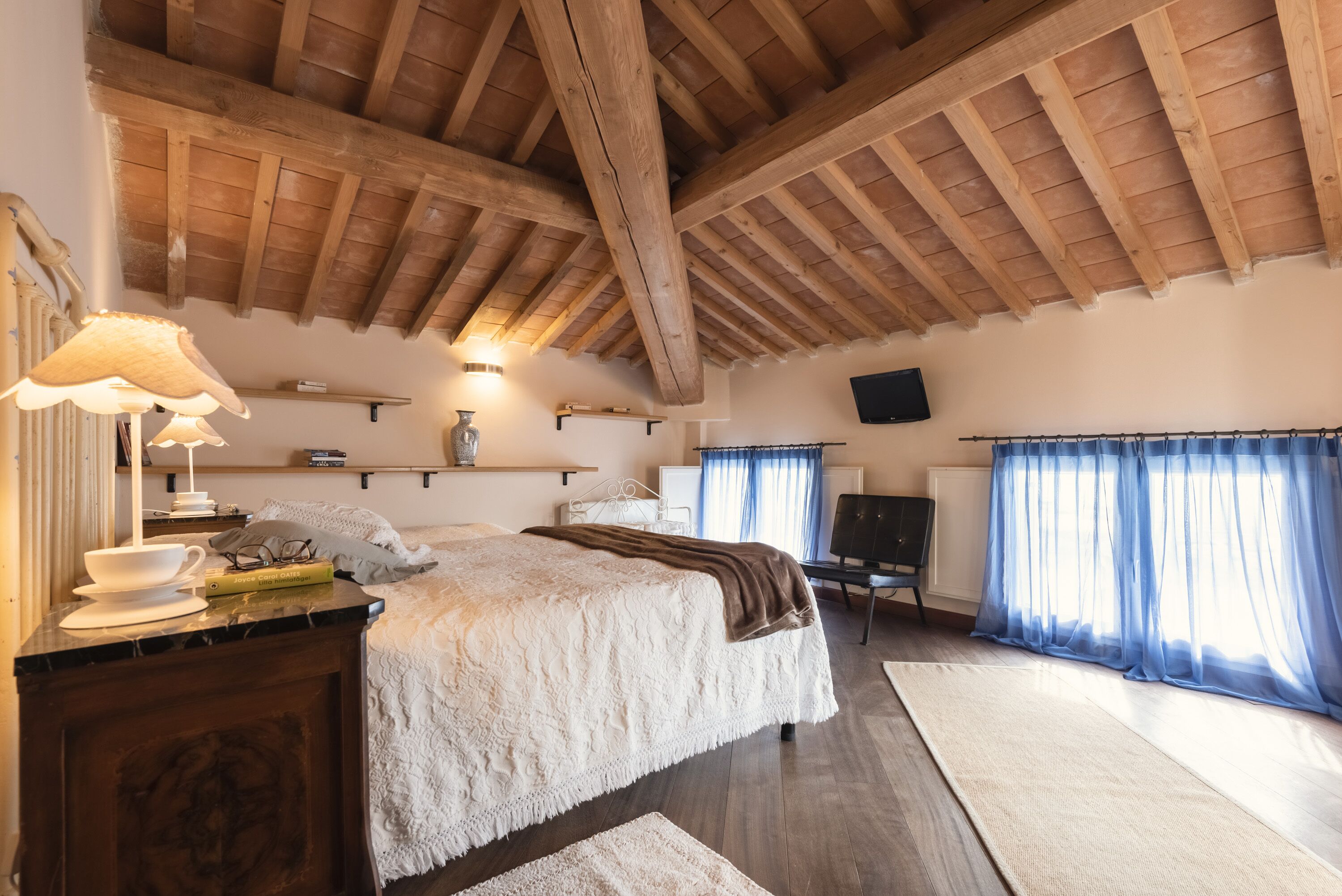 Quarto com teto de madeira, cama com colcha branca, mesa de cabeceira com abajur e janela com cortinas azuis.