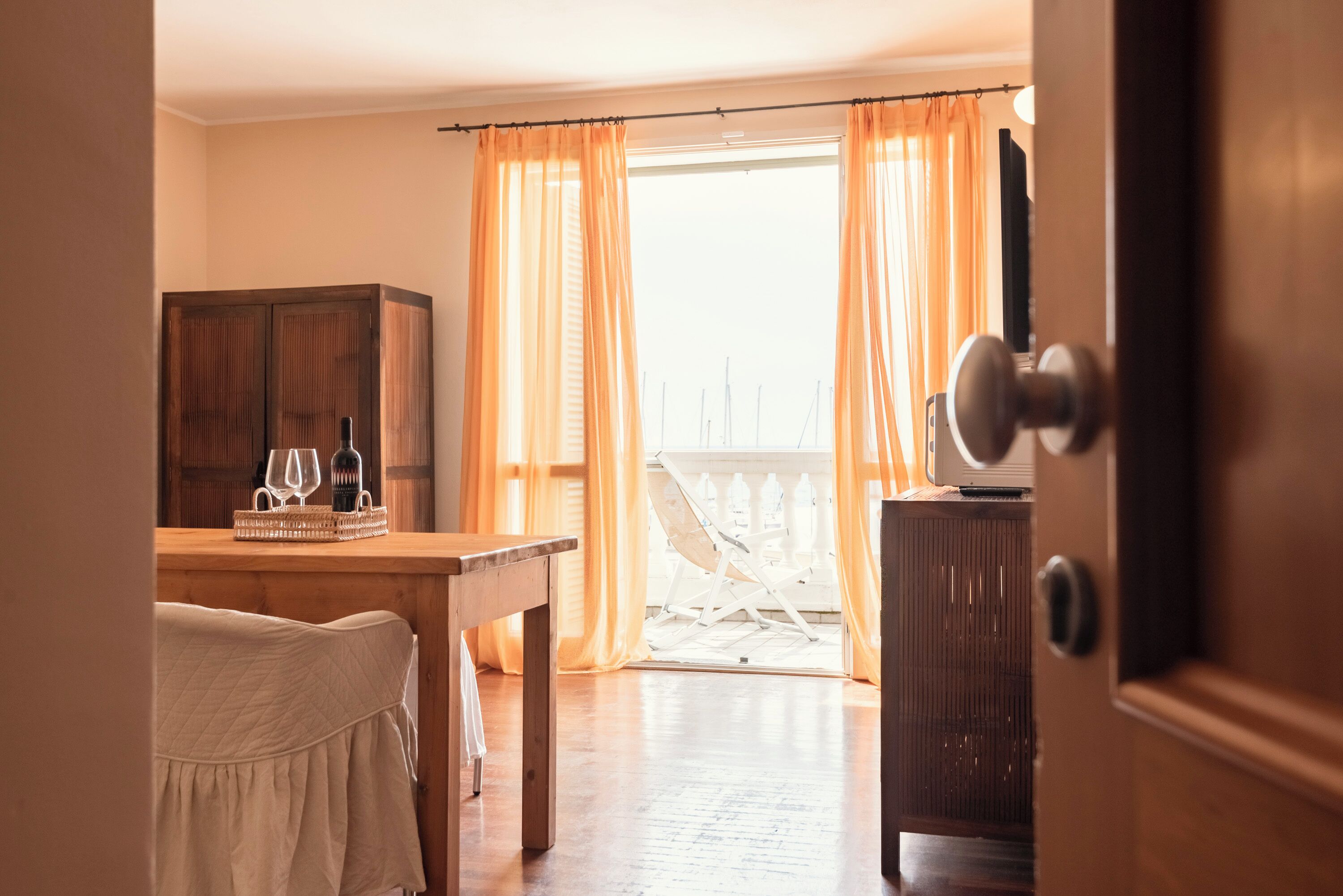 A room with a wooden table, a chair, and a wardrobe. There is a balcony with a white railing and orange curtains.