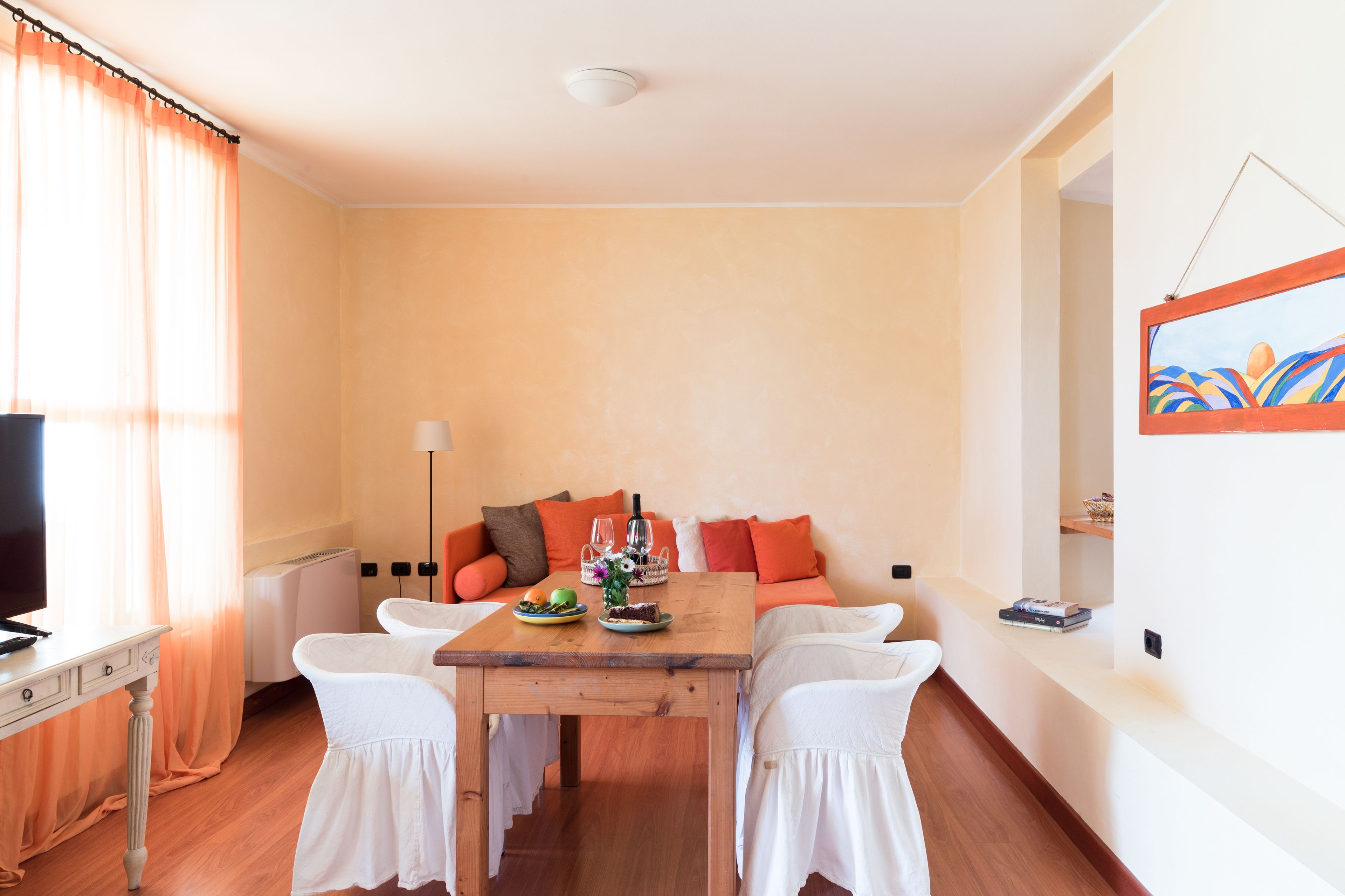 Ein Zimmer mit einem Holztisch, weißen Stühlen, einer Couch mit orangefarbenen Kissen, einem Fernseher und einem Bild an der Wand.