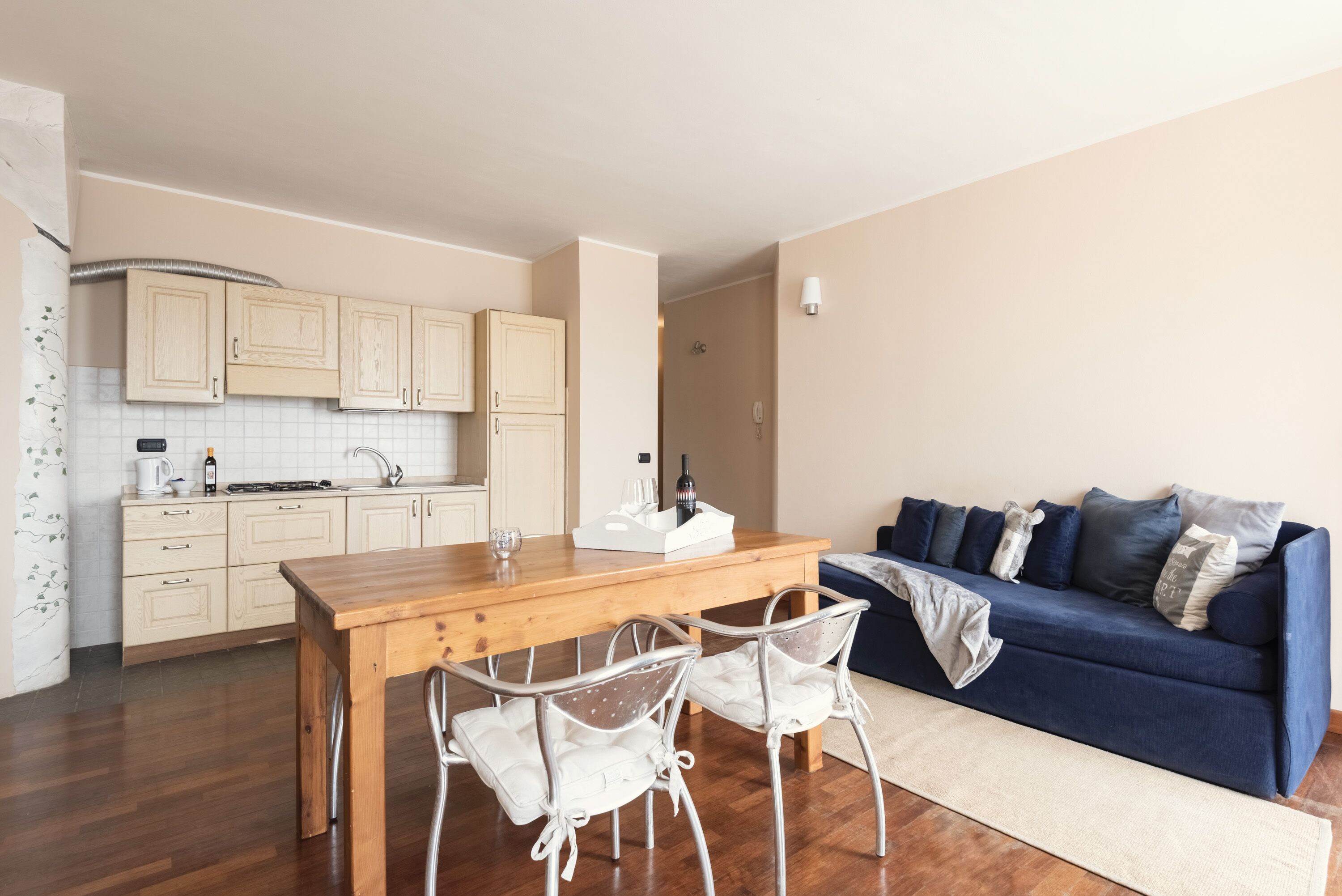 A compact living space with a wooden dining table, two white chairs, a blue sofa, and a kitchen area with light-colored cabinets and a white backsplash.