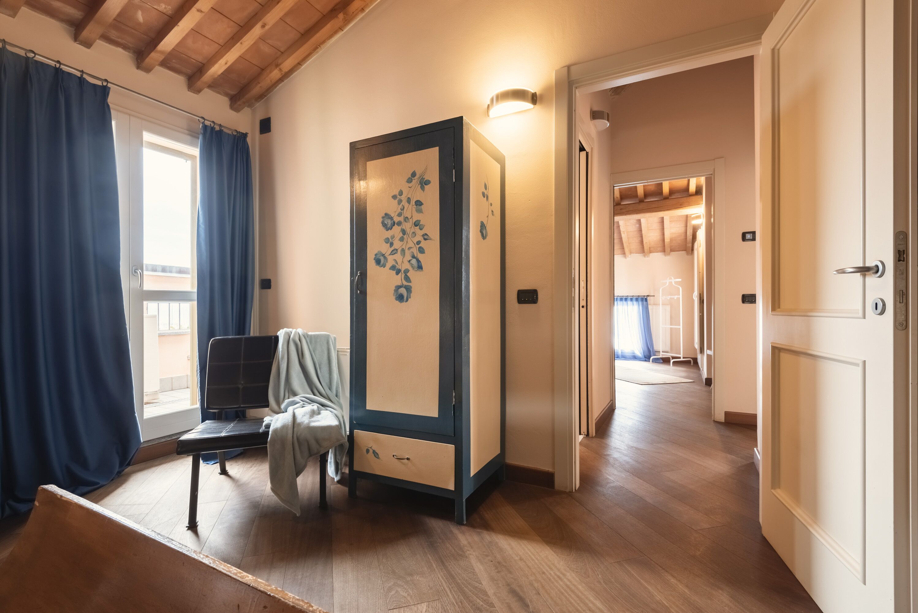 A room with a wooden floor, a bench, a wardrobe with floral designs, and a door leading to another room.