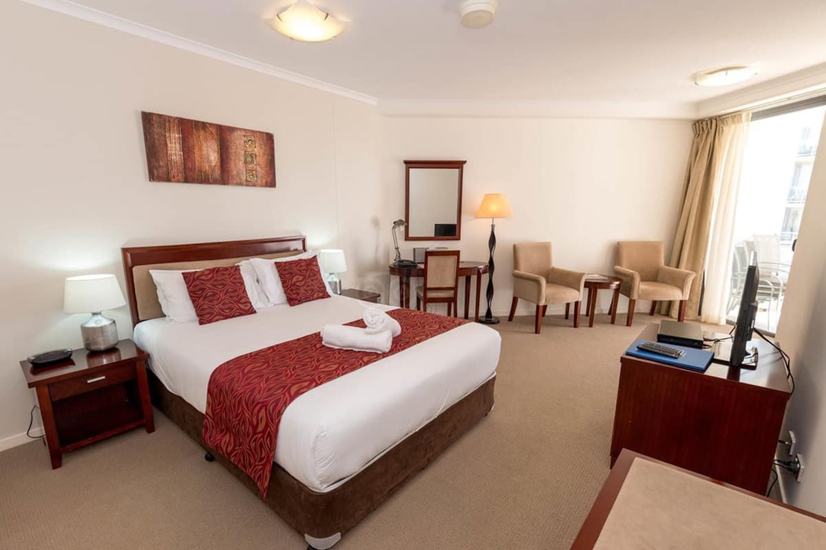 A hotel room with a large bed, a desk, two chairs, a TV, and a window with curtains.