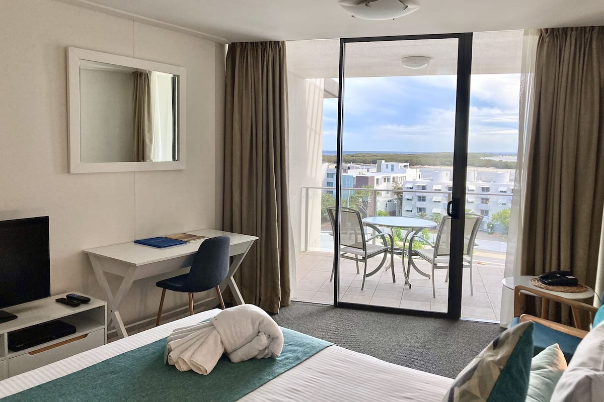 A hotel room with a bed, a desk, a chair, a TV, and a balcony with a view of buildings.
