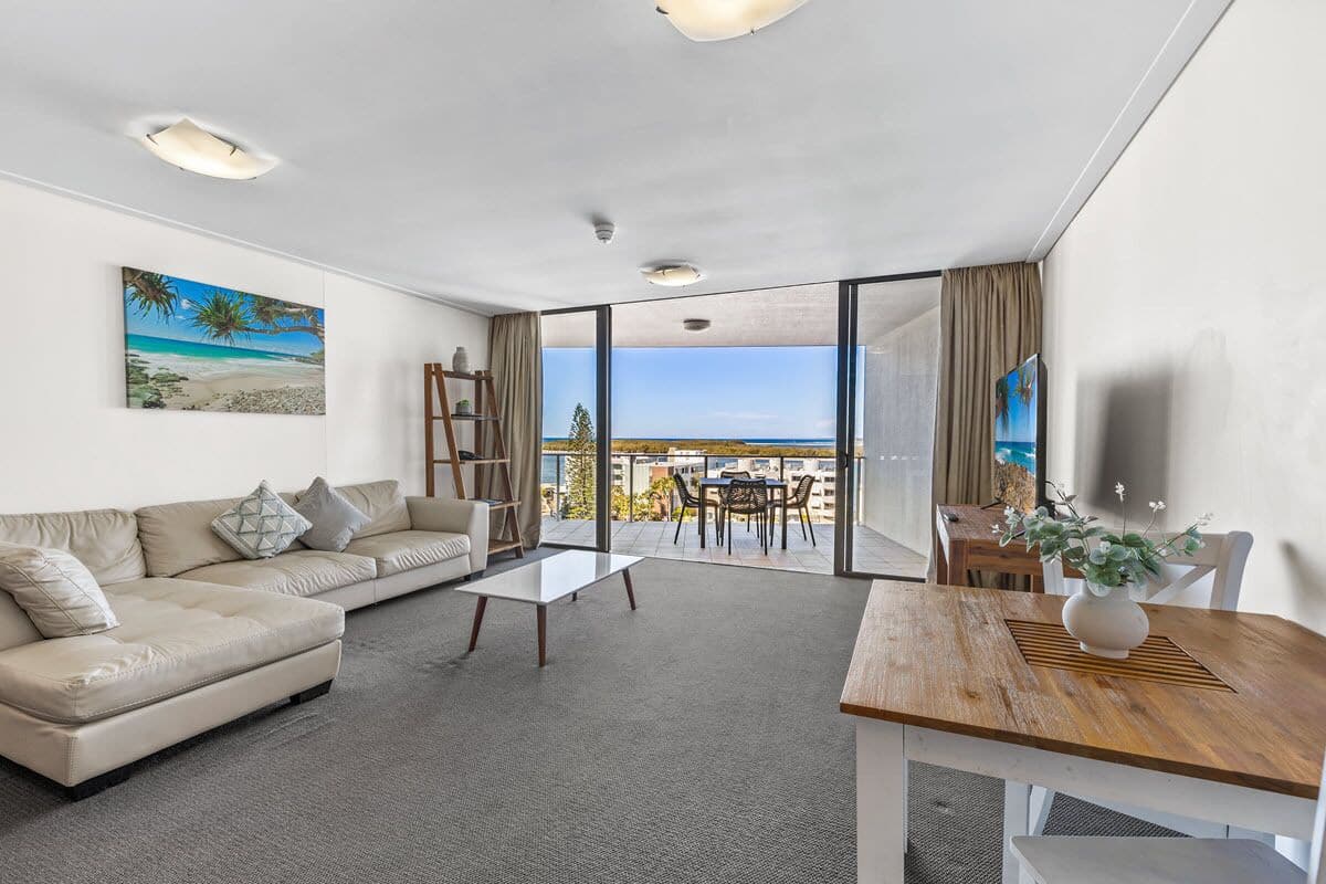 A modern living room with a sofa, coffee table, and dining table, a balcony with a view, and a painting on the wall.