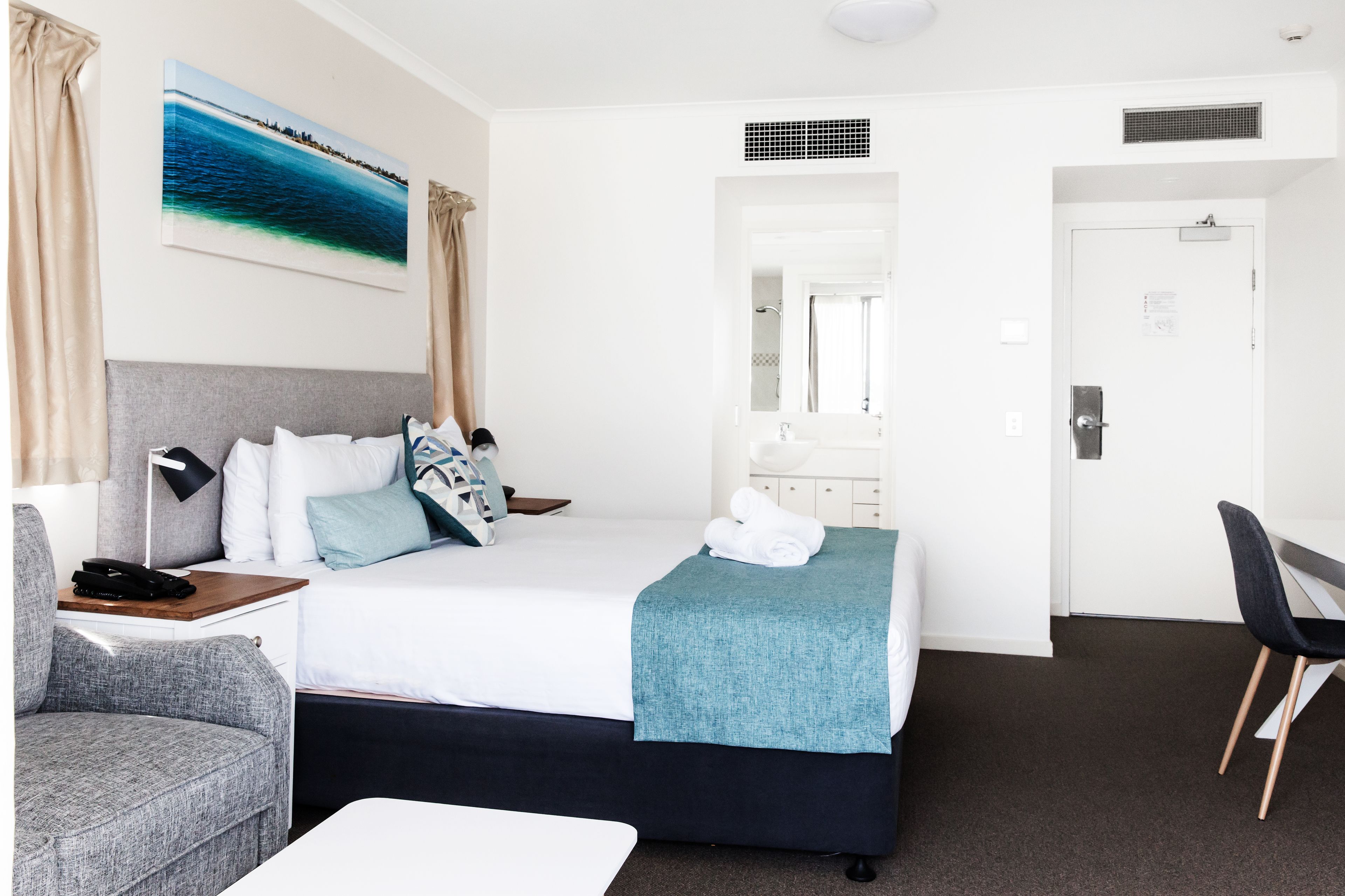 Habitación de hotel con cama, escritorio, silla, lámpara de escritorio, mesita de noche, ventana con cortinas y un cuadro en la pared.