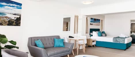 Una habitación de hotel moderna con un sofá gris, una mesita redonda pequeña, una cama con ropa de cama color turquesa y un ventanal con vista a la playa.