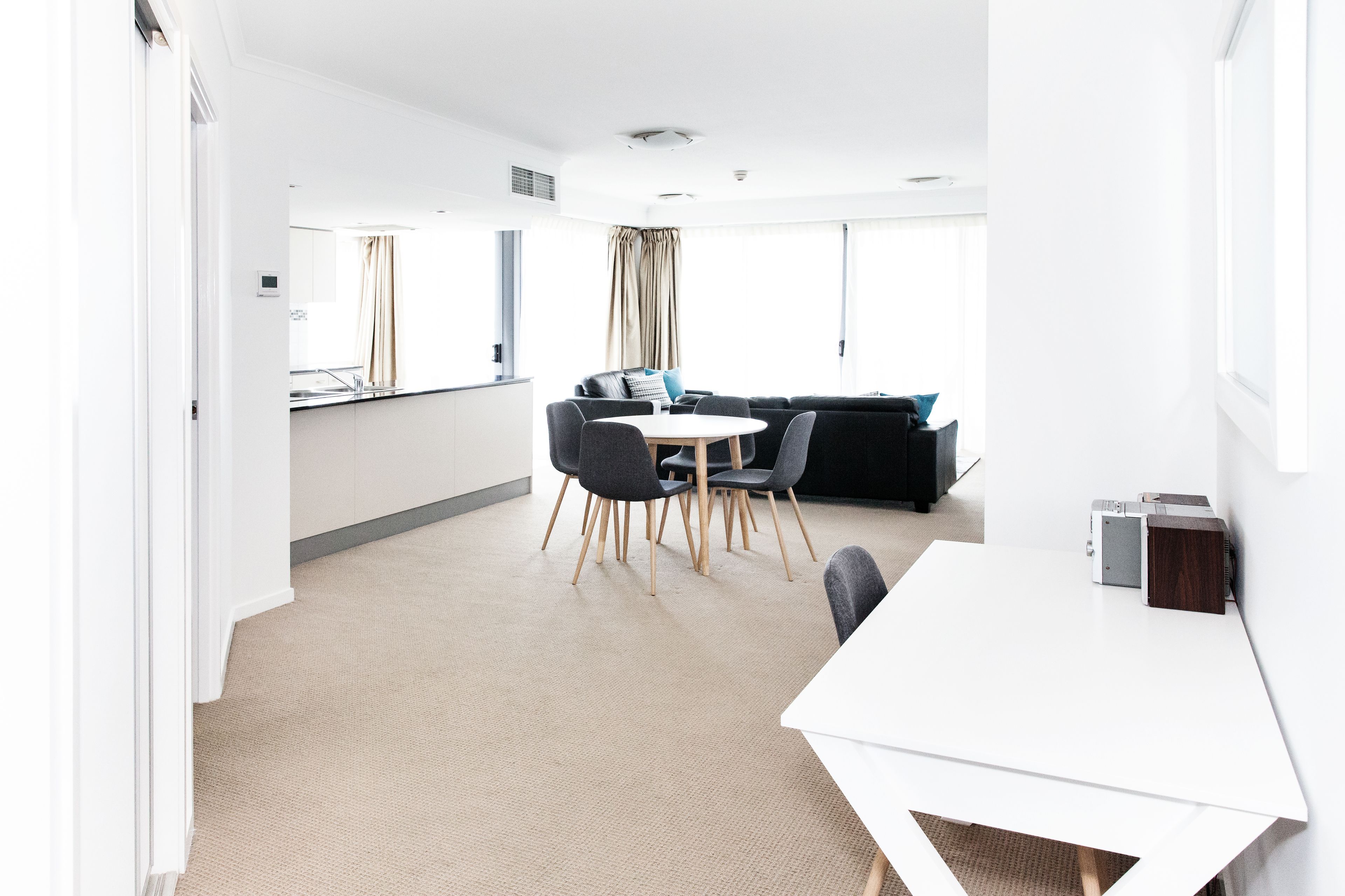 A modern hotel room with a white bed, a large window, a sofa, a dining table with chairs, and a kitchen area.