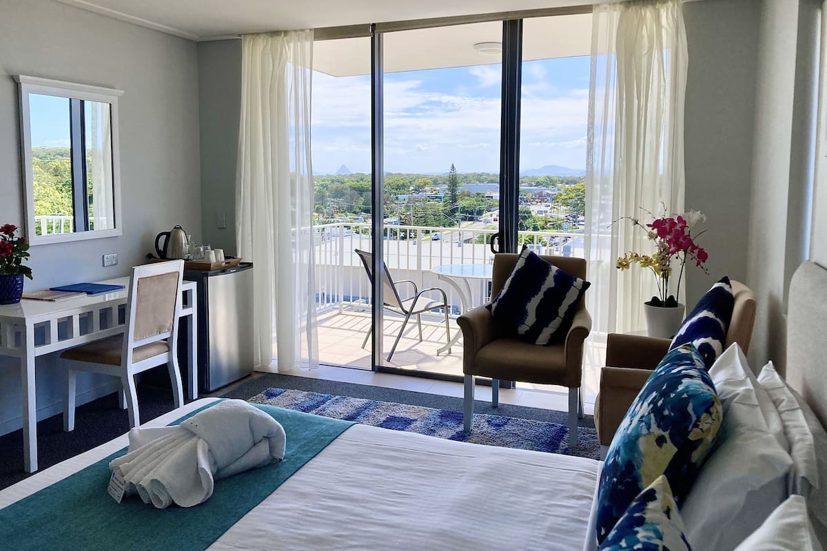 A hotel room with a bed, a desk, a chair, a balcony with a view, and a window with curtains.