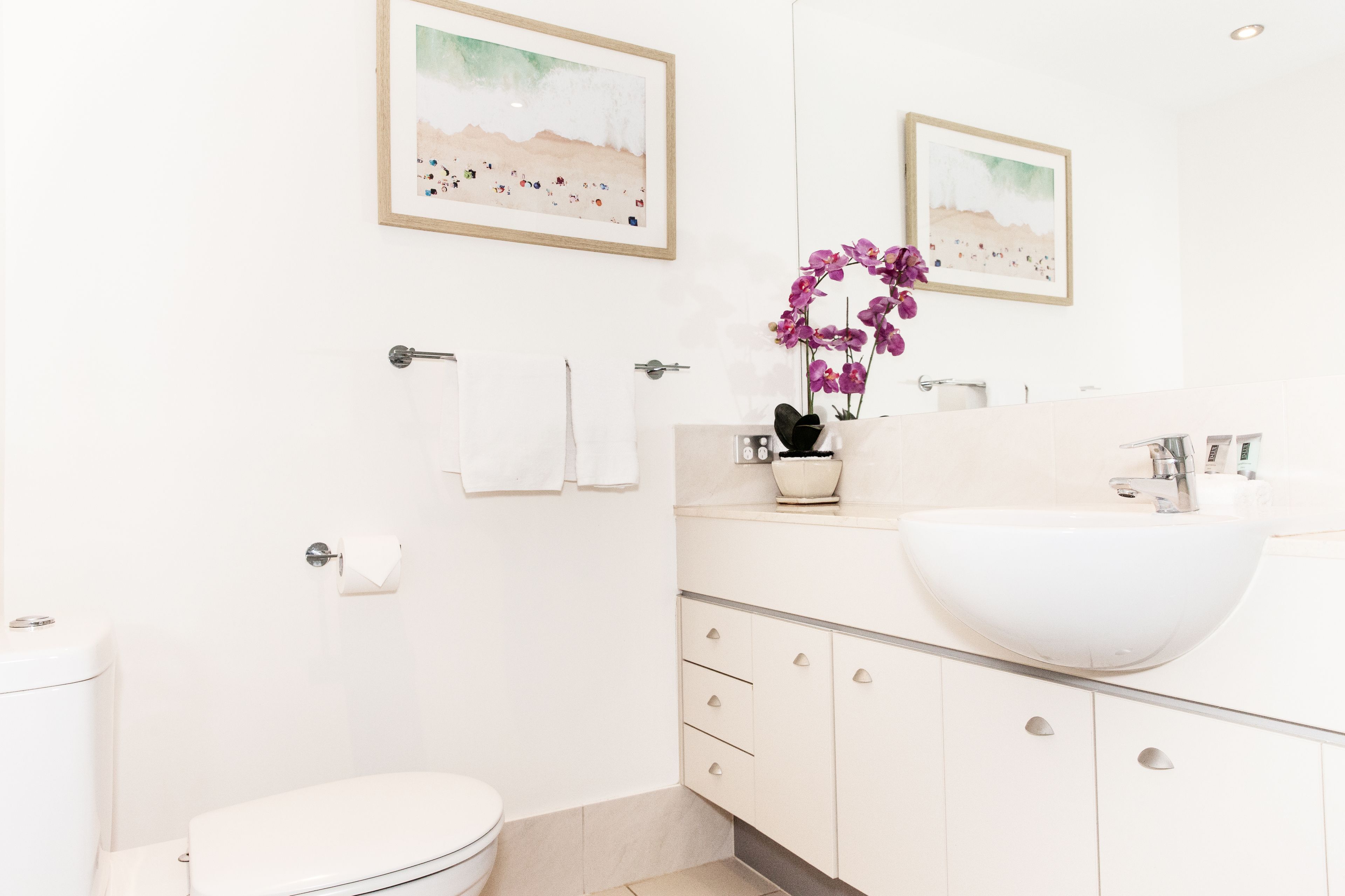 Un baño moderno con lavamanos, inodoro y tocador blancos. Hay dos cuadros en la pared, un florero con flores moradas y un perchero con dos toallas.